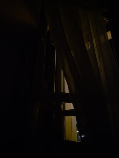 The image depicts a dimly lit interior looking out of a window at night. The foreground is dominated by dark shadows, suggesting the viewer is inside a room. Partially obscuring the view is a light-colored curtain, its fabric creating folds and casting further shadows. Beyond the curtain, a window frame is visible, divided into sections by vertical and horizontal mullions. Through the window, faint exterior lights can be seen, indicating an urban environment. The overall impression is one of nighttime and indoor privacy, with minimal external details discernible. The location is identified as Montrouge, France.