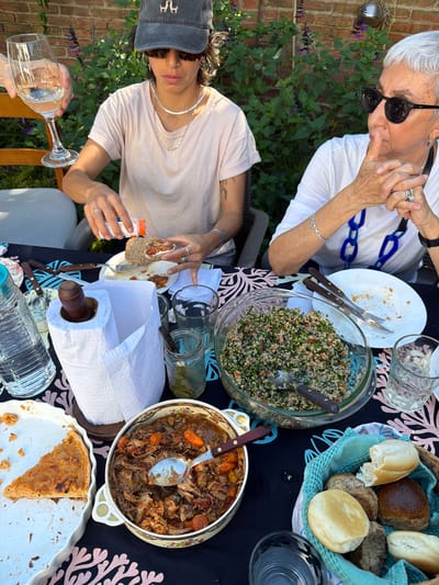 The image shows an outdoor gathering of two people at a table laden with food and drinks. The person in the foreground holds a glass of wine and appears to be preparing a sandwich, adding a condiment from a white container. The person in the background is older, wearing sunglasses and a white top with a blue chain pattern. On the table, there is a salad bowl, a basket with assorted bread, a dish of stew-like meat, and a partially eaten quiche or pie. The setting suggests a backyard or garden with brick walls and lush greenery. Location: Tolosa, Argentina is not apparent from the image.