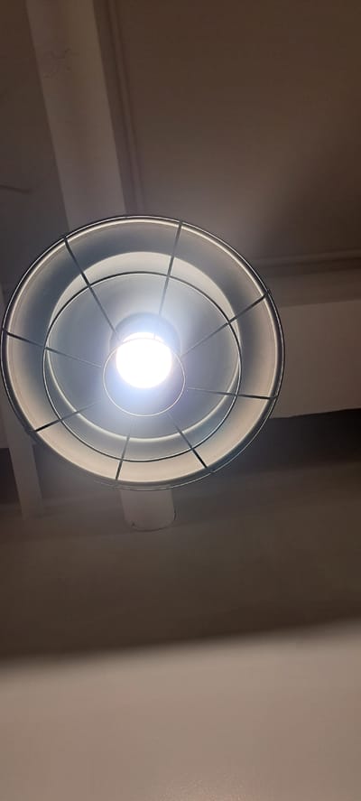 The image shows a close-up, low-angle shot of a hanging light fixture, likely an industrial-style pendant lamp with a metal cage. The light bulb within the fixture is illuminated and emitting a bright, white light. The lamp is suspended from a ceiling which appears to be painted a neutral, light color, possibly white or off-white. There are some faint marks or shadows on the ceiling. The background is largely composed of the ceiling and upper walls, which are also painted in muted tones, creating a simple and uncluttered environment. The scene is devoid of people or any other discernible objects besides the light fixture and the ceiling. The overall impression is one of an indoor space, likely a room or hallway, illuminated by this single fixture.

Based on the provided metadata, this image was taken in Mandaluyong City, Philippines. The location suggests an indoor setting within the city, possibly a residential, commercial, or public space. The specific details of the image do not provide further clues to pinpoint an exact location within Mandaluyong City.