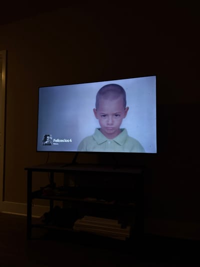 A television displays a picture of a young, possibly Latino boy wearing a light green collared shirt. The television is mounted on a wall above a dark metal stand with multiple shelves. The surrounding environment is dimly lit, making details in the room challenging to discern. The image displayed on the screen includes the name "Felices los 4" alongside a thumbnail image, indicating it may be a music video or streaming content.