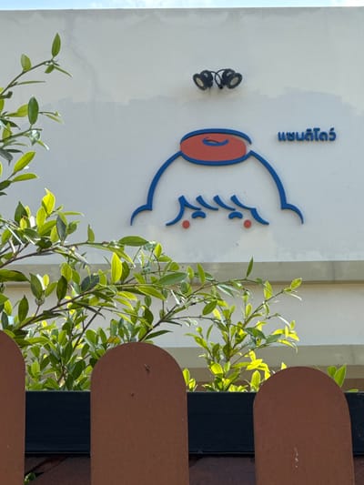 The image displays an exterior daytime scene in San Phra Net, Thailand, focusing on a white building wall with distinct signage, partially obscured by vegetation and a fence. The primary subject is a stylized logo on the wall, featuring a blue outline of a head-like figure, an orange-red circular element with a blue curve on top, and two small red dots within the face area. Adjacent to this logo, the Thai script "แซนดิโก๋" (Sandigo) is rendered in blue. Above the logo, two dark, circular fixtures, possibly lights or cameras, are mounted on the wall. The lower portion of the frame is dominated by a brown, arched, fence-like structure, behind which lush green foliage is visible, illuminated by direct sunlight. The wall exhibits minor cracks. No individuals are present.
