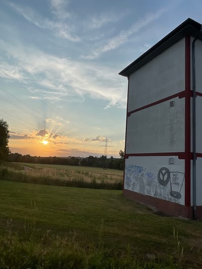 A late afternoon scene captures a residential building with graffiti on its white facade, marked with red trim and a dark roof. The building is situated in the rural outskirts of Rückershausen, Germany, overlooking a field of dry grasses and vegetation. In the background, a power line pylon stands tall against a sky filled with a setting sun. The sun's rays break through scattered clouds, casting a warm glow over the landscape and creating dramatic light shafts. The area appears to be a field with some trees and distant structures.