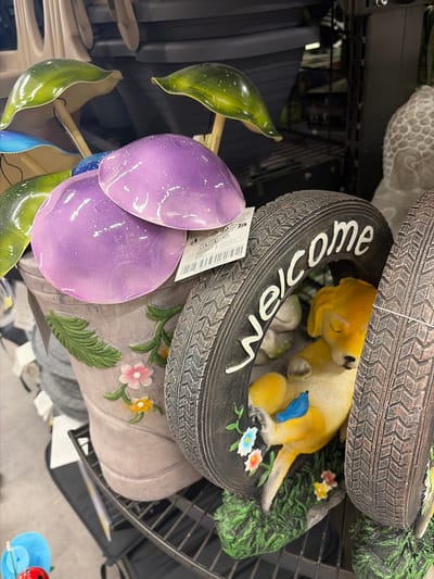 This is a close-up, eye-level, indoor shot of garden decorations displayed on a black metal shelf. In the foreground, a light gray planter shaped like a boot features embossed green leaves and pink and yellow flowers. Resting on the edge of the planter and extending out are large, shiny purple mushroom caps with white speckles, and a green mushroom cap with a glossy finish. To the right of the planter is a tire-shaped decoration with the word "welcome" in white lettering. Inside the tire, a yellow dog figurine is sleeping, with a small blue bird perched on its hindquarters. The dog rests on a green, grassy base with a few small flowers. The background shows other shelves with more garden decor and dark plastic containers. The image is sharp and well-lit, focusing on the whimsical garden figures. This scene is not located in Würzburg, Germany.