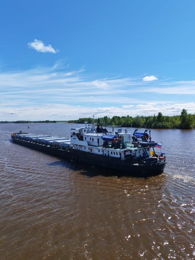 The image depicts a large cargo barge, identified as "KOSINO" in Cyrillic script, traversing a wide river. The barge is loaded with numerous gray cargo containers. The body of water is a muddy brown color. Lush green trees line the riverbank in the background. The sky is bright blue with scattered white clouds. The overall setting suggests a location within Russia, specifically near Kargulino given the information. The Russian flag is visible on the stern of the barge.
