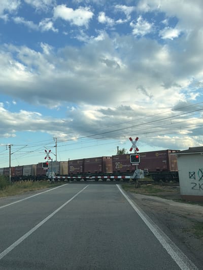 In Luhyňa, Slovakia, a train of freight containers moves across a railway crossing. The crossing has a red signal light illuminated, indicating that it is unsafe to cross. The train consists of several dark red containers with "ZTS" and "innofreight" branding. On the right side of the image, a white building with graffiti is partially visible. The sky above is filled with dramatic, cumulus clouds. The foreground shows a paved road with white lane markings.