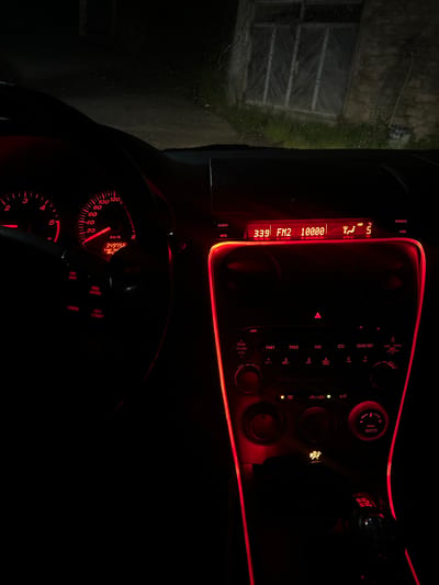 The image depicts the interior of a car, focusing on the dashboard and steering wheel. The lighting is predominantly red, illuminating the car's instruments and controls. The radio display shows "3:39 FM2 10000." The instrument panel displays a speed of approximately 15 km/h and an odometer reading of 349756. The steering wheel includes cruise control buttons. Outside the car window, a dimly lit building or structure is visible through a rain-streaked window. There is no information to determine the location as the city of Kribul, Bulgaria.