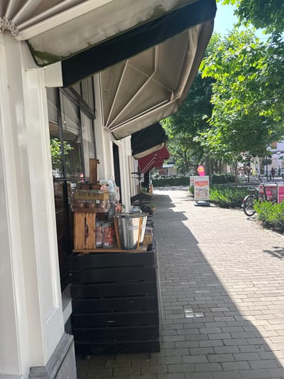 The image depicts a street scene in Zwolle, Netherlands, on a sunny day. The perspective is from a ground-level viewpoint, looking slightly upwards and to the right. In the foreground, a white window frame and part of a building facade are visible. A retractable awning, partially open and a muted green color, extends over a window.

Beyond the building, a paved sidewalk stretches into the distance, lined with trees on the left, casting dappled shadows. Along the sidewalk, there's a display of goods, including wooden crates filled with items. One crate has the text "WATCH ME GROW" printed on it. A silver bucket sits atop the crates. Further down the street, a bicycle is parked, and there are signs visible, one of which reads "Vandaag 20% KORTING," indicating a 20% discount is available today. Other signs and shop fronts suggest a commercial area. In the background, buildings and a few indistinct figures can be seen, hinting at typical street activity. The bright sunlight and clear shadows suggest it is midday. The overall impression is a vibrant, daytime scene in a Dutch town.