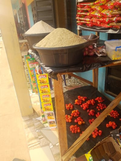 The image captures an outdoor market stall, brightly lit, likely under sunlight, suggesting it's daytime. The stall features a variety of food items for sale, displayed on wooden structures.
In the foreground and center, two large, dark-colored plastic tubs are prominently displayed, filled with grains. The tub in the front contains a large mound of light brown or beige grains, likely parboiled rice. Slightly behind and to its left, another similar tub holds a mound of white grains, presumably white rice. Both tubs are placed on a wooden table or stand, which is covered with a black tarp or plastic sheet.
To the right of the front grain tub, on the same table, there's a small pile of red onions and a clear plastic storage container with a blue lid, possibly holding smaller packaged goods. Behind the rice tubs, further back and elevated, a stack of packaged instant noodles or similar food items in red and yellow wrappers is visible.
To the left of the grain display, a narrow, vertical rack holds numerous small, yellow packets. Each packet clearly shows the brand name "ARSOR" and an image of a roasted chicken, indicating they are likely chicken seasoning cubes or powder. The text "CHICKEN CUBE" or "CHICKEN SEASONING" is partially visible below the brand name.
Below the main table, on a lower tier or shelf, also covered with a black tarp, are several small clusters of vibrant red and orange-red peppers, likely scotch bonnets or similar hot pepper varieties. These peppers are irregularly shaped and appear fresh.
The ground beneath the stall is tiled with light-colored, possibly beige or white, patterned tiles. To the far left, a light-colored, possibly beige or yellow, building wall is visible, contributing to the outdoor setting. In the background, hints of other structures with blue and darker tones suggest a street or market area. To the bottom right, a brown cardboard box containing some dark, rectangular items, possibly chocolate or soap bars, is partially visible. The overall scene depicts a bustling local market environment with various essential food items.