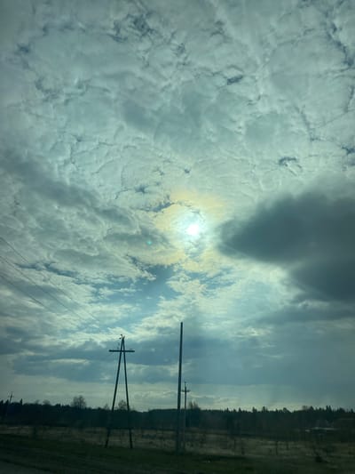 The image shows a daytime scene in what appears to be a rural or semi-rural area. The sky is predominantly cloudy, with patches of blue visible. The sun is obscured by the clouds but still emits a bright glow. Power lines and telephone poles are visible, stretching across the frame, indicating the presence of infrastructure. A treeline or forest is present in the distance. It is not possible to definitively confirm the location as Malaya Bod'ya, Russia, based solely on visual information.