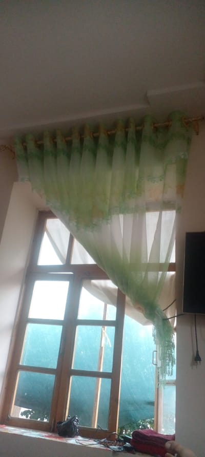 An interior shot shows a window with a light green sheer curtain that has a decorative floral pattern along the top and bottom edges. The curtain is hung on a golden rod. Outside the window, the glass is tinted a bluish-green, and the background appears to be foliage. On the windowsill below the window, there are several objects, including a black plastic bag, wires, and a stack of folded red fabric. To the right of the window, a black television screen is visible, mounted on a wall. The lighting in the room is dim, with light coming from the window. The image's composition suggests it was taken from a slightly upward angle. The scene is set in Parkent, Uzbekistan.