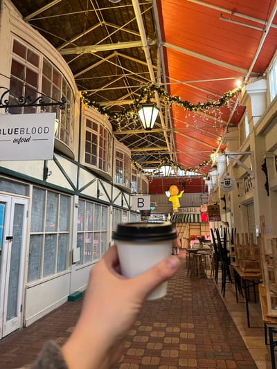 The image captures an indoor passage within Oxford's Covered Market, United Kingdom. In the foreground, a fair-skinned hand holds a white paper coffee cup with a black lid, oriented slightly right of center. The passage features a vaulted ceiling structure with exposed metal beams and alternating reddish-orange and dark paneling. Overhead, strings of green garland with white lights and icicle-like light strands are draped, accompanied by hanging, dark-framed lantern lights.
On the left side of the passage, a row of white-framed shopfronts is visible, characterized by large, multi-pane windows and distinctive curved bay windows on their upper sections. A white rectangular sign suspended from a black bracket reads "BLUEBLOOD oxford." A square sign with the letter "B" is visible further down this side.
On the right side of the passage, a narrower aisle shows multiple tables and chairs, some stacked, suggesting either pre-opening or post-closing conditions. Shop signs are present, including a partially visible "BONNERS" sign and a round sign labeled "Gerry's Cellar Door." A large, illuminated yellow anthropomorphic figure, possibly a pig, hangs from the ceiling in the distance. A blurry, distant figure in striped attire is faintly visible on the right. The floor of the passage is paved with light and reddish-brown bricks arranged in a basket-weave pattern.