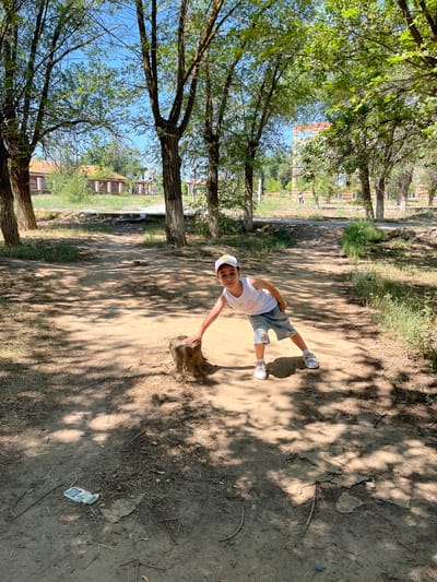 A young boy wearing a white tank top, denim shorts with rips, a white baseball cap, and white sneakers is posing in a park in Ikryanoye, Russia. He is leaning down, with his right hand resting on a tree stump in the foreground. The park features a dirt path, scattered dry grass and leaves, and several tall trees casting shadows on the ground. In the background, there are buildings with reddish roofs and some vegetation. The sky is clear and blue.