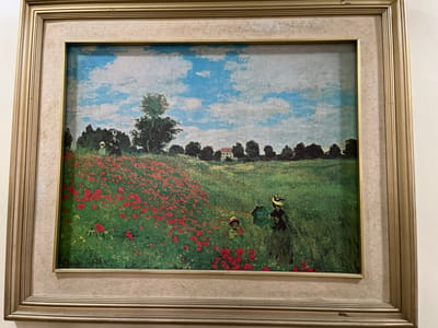 The image is a framed painting of a pastoral scene. The painting depicts a lush green field dotted with vibrant red poppies in the foreground and middle ground. In the distance, a line of trees forms a dark silhouette against a bright blue sky with scattered white clouds. A small, white house with a red roof is visible nestled amongst the trees.

Two figures are present in the field. To the right, a woman in a dark dress and a wide-brimmed hat stands facing left, seemingly looking towards the poppies. In the middle of the poppy field, a child wearing a hat is peeking out from among the flowers. The overall impression is one of a serene, sunny day in the countryside. The light suggests it is likely daytime, with the sun high in the sky.

The painting is housed in a textured, light-colored mat and a golden, multi-layered frame. There appears to be a signature at the bottom right of the painting, though it is partially obscured and difficult to read definitively. The location context provided is Quezon City, Philippines, though the painting itself depicts a European landscape, likely painted by an artist inspired by French Impressionism, given the subject matter and style.