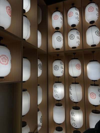 The image displays an indoor wall decorated with numerous illuminated Japanese paper lanterns, known as chochin, arranged in a grid pattern. The lanterns are cylindrical, white, and internally lit, casting a soft glow. Various designs are present: some lanterns feature a red stylized floral emblem, while others bear a gray or black Japanese family crest (mon). Many lanterns also display black Japanese characters that translate to "Teppanyaki" (鉄板焼). These lanterns are mounted or suspended within compartments formed by light-colored wooden panels, which serve as the background. No individuals or specific actions are visible in the scene, which is part of a Japanese establishment in Clamart, France.