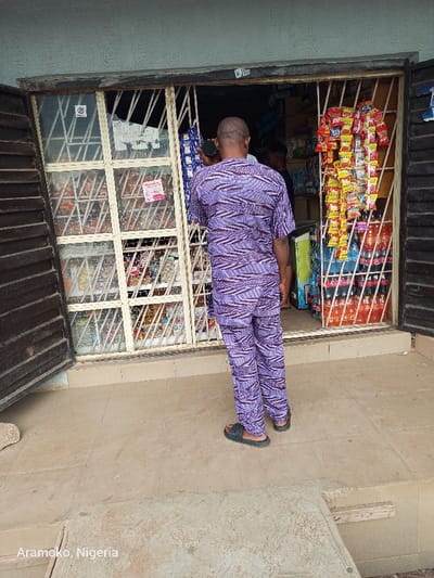 The image shows a man wearing a purple and brown patterned shirt and pants standing in front of a store with a metal grate door. The store is located at coordinates (latitude: 7.839, longitude: 4.484) in Ilobu, Nigeria. Inside the store, shelves are stocked with various goods, including bags of chips, bottles of soda, and other items. Another person is partially visible in the background. The sidewalk in front of the store is made of tan-colored concrete tiles.