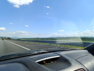 Captured from within a moving vehicle, the image presents a highway landscape in Vakarel, Bulgaria, under a bright, blue sky with scattered clouds. The vehicle's dashboard and windshield frame the view. A multi-lane highway stretches ahead, separated by a metallic guardrail. Beyond the guardrail, a verdant expanse of fields and forests extends towards the horizon, where distant mountain peaks capped with snow are visible. The windshield shows light reflections, indicating sunlight. Another vehicle is seen in the distance on the highway.