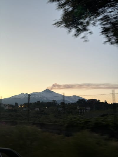 The image, likely taken from a moving vehicle, presents a landscape with a mountain in the background. The sky is a pale, gradient color, shifting from a light gray to a yellowish hue near the horizon. Smoke is visible above the mountain. A blurred tree branch is in the upper left corner. In the middle distance, a fence runs along the edge of a green area with some buildings. The right side of the image is blurred, possibly due to the movement of the vehicle. The location might be near Acireale, Italy, given the presence of Mount Etna.