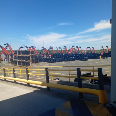 The image shows an outdoor lot filled with numerous red and black telescopic handlers, commonly known as telehandlers. These machines, manufactured by Manitou, are lined up in rows, with their booms extended at various angles. Many of the telehandlers have black buckets attached to their front ends, suggesting they are either ready for operation or storage. In the foreground, there are stacks of black containers or pallets. A yellow barrier with black posts runs across the bottom of the frame, separating the lot from a paved area. The sky is a clear blue with a few wispy clouds. The scene appears to be a storage or staging area for this type of heavy machinery. The location is identified as Ancenis, France.
