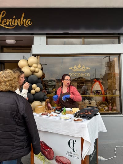 An outdoor scene captured in front of the "Leninha" confectionery in Felgueiras, Portugal, depicts a food tasting event.
A female vendor, with reddish-brown hair and glasses, wearing a salmon-colored shirt, brown apron, and blue gloves, stands behind a white-draped table. She gestures with her right hand while addressing two female customers. The customer in the foreground, seen from the back, wears a black quilted jacket. The second customer, slightly behind her, with brown hair, is tasting a food sample from a toothpick.
The table features a variety of food items: a leg of cured meat on a wooden stand, multiple plates with sliced meats and cheese samples served with toothpicks, a block of cheese, and a small green bottle. On the right side of the table, an electric griddle contains cooking slices of meat. The front of the table is adorned with a banner promoting "Quinta dos Fumeiros," showcasing images of smoked meats. A white power strip with a cable is visible on the ground to the right, powering the griddle.
To the left of the table, a beige and black balloon cluster stands. Behind the vendor and the table is the storefront window of "DOCINHOS Helena Gonçalves - Docaria Tradicional," displaying an oversized Easter egg decoration and ceramic bunny mugs, indicating an Easter theme. The store's primary sign, "Leninha," in gold script, is mounted on the black fascia above the window. The sky, reflected in the window, appears overcast.