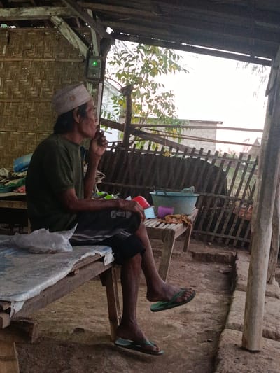 A full shot shows a middle-aged man smoking a cigarette while sitting on a wooden bench under a thatched roof structure in Lamongan, Indonesia. He wears a green t-shirt, black shorts, and a white skullcap. To his right, a teal-colored tub rests on a wooden table with several other objects. The bench is covered with a white sheet and a plastic bag is on the left side. The man is in the process of exhaling smoke, his eyes closed. The background is made of woven bamboo and a wooden fence. The lighting suggests it is late afternoon, with the sky visible through the trees in the background.