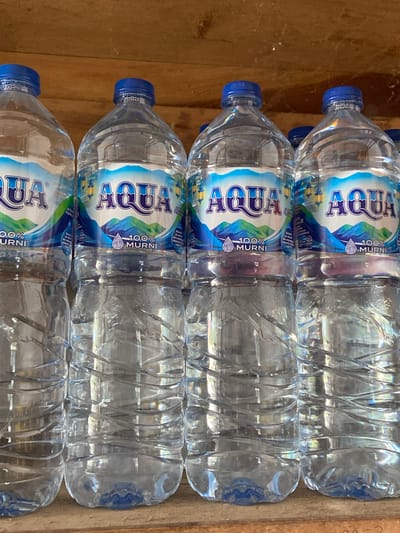 A close-up, eye-level shot shows a row of six identical, clear plastic bottles of AQUA brand bottled water lined up on a light brown wooden surface. The bottles have blue caps and labels featuring a blue and green mountain landscape with a crescent moon and lanterns. Each bottle has "AQUA" written in prominent white letters on a blue background. Below this, "100% MURNI" is written in white text with a circular water droplet logo. The bottles are filled with clear water, and the lower portions show the characteristic ridging of plastic water bottles. The background consists of the textured wooden surface on which the bottles rest. The scene is likely from Sekar Mawar, Indonesia, a location known for its natural beauty and potentially its water sources.