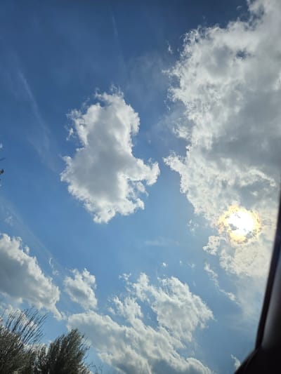 The image shows a view of the sky with white clouds and a bright sun. The sky is blue. The photograph was taken from a low angle, possibly from inside a vehicle. A section of what appears to be a vehicle frame is visible in the upper portion of the image. The location in the city of Koilovtsi, Bulgaria cannot be verified from the image content.