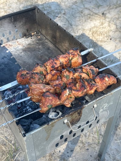 The scene depicts a charcoal grill with cooked meat skewers. The grill is rectangular, metallic, and shows signs of wear with soot and residue. Inside, there are charred coals, indicating recent use. Four metal skewers laden with cooked meat are positioned across the top of the grill. The meat appears browned, slightly charred, and sprinkled with green herbs. The grill rests on a surface that appears to be sandy or covered in dry grass. The setting may be an outdoor area on the outskirts of a city. There is no indication that the image was taken in or around the city of Zīvartiņš, Latvia. The image provides no information as to people, interactions, or actions other than cooking. The image is a still photograph with no people visible.