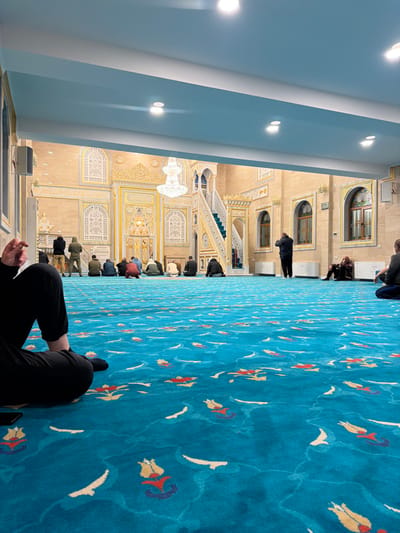 This image captures the interior of a mosque in Breznitsa, Bulgaria, viewed from a low angle, looking towards the front. The floor is covered by a vibrant turquoise blue carpet patterned with stylized red, yellow, and white floral and wave motifs. The walls are light tan, extensively adorned with intricate gold-colored relief decorations, including geometric designs, arched panels, and what appears to be calligraphy. A prominent central archway, likely a mihrab, is visible at the rear. To its right stands a white staircase with ornate balustrades and a green carpeted path, likely a minbar. Arched windows with wooden frames are visible on the right wall. The ceiling is light blue with white recessed beams and multiple embedded circular downlights, complementing the illumination from a large, multi-tiered crystal chandelier hanging in the mid-background.
In the foreground, the lower legs and torso of an individual wearing black pants and socks are visible, seated on the carpet. A dark mobile phone rests on the carpet near their left leg. In the mid-background, approximately 8-10 adult males are observed from behind, arranged in rows on the carpet, engaged in prayer; most are in a kneeling or prostrating position. To their right, one adult male stands facing the back wall with hands raised in a prayer posture. Further right, another adult male is seated on the carpet, leaning against the wall with his head down. On the far left, two other adult males are standing near the wall, also facing the back. A white rectangular speaker is mounted on the left wall, and white ventilation units are situated along the base of the right wall.