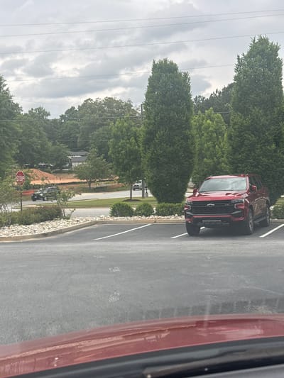 The image shows a red Chevrolet Colorado pickup truck parked in a parking lot next to a grassy area with trees in Stone Mountain, Georgia, United States. In the background there are more trees and a construction site. A stop sign is visible near the construction area. Another red vehicle is partially visible on the right edge of the frame, suggesting the photo was taken from inside the vehicle. The sky is overcast.