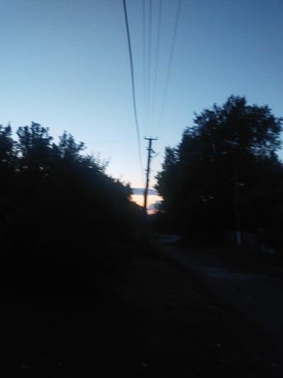 The image depicts a rural scene in Sosnivka, Ukraine, at dusk. Silhouetted trees line a dirt road that curves into the distance. A single utility pole stands prominently to the left of the road, with several wires extending upwards and across the frame. The sky is a gradient of pale blue and light orange, indicating the setting sun. The overall mood is serene and quiet, typical of a rural evening. The low light conditions and the motion blur suggest the photograph was taken either at night or while moving. There is no visible text in the image.