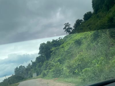 The image depicts a view from a moving vehicle on a road, likely in a tropical region. The foreground shows a portion of the asphalt road, with a gravel shoulder to the right. A steep, densely vegetated hillside rises from the road on the right side of the frame. The hillside is covered in lush green foliage, including various bushes, trees, and tall grasses. One prominent tree with a distinctive silhouette stands out on the upper part of the slope.

In the distance, to the left of the frame, the landscape appears flatter, with more trees and some power lines visible. The sky is overcast with heavy, dark gray clouds, suggesting impending rain or a generally gloomy day. The lighting is diffused, contributing to the somber mood of the scene.

The overall scene is one of a natural, undeveloped landscape, possibly a rural area in Nigeria, as indicated by the mention of Junyura. There are no people or specific objects that draw immediate attention other than the natural elements and the power lines. The image seems to capture a moment of travel through a green, hilly terrain under a cloudy sky. The time of day is unclear due to the overcast conditions, but the diffused light suggests it is daytime.