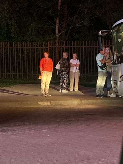 The image, taken in Terespol, Poland, depicts a group of four individuals standing outdoors at dusk. The scene appears to be near a fence and a paved area, possibly a sidewalk or parking lot. The lighting suggests it is evening, with artificial light sources illuminating the subjects.
From top to bottom, the individuals are:
The first person, partially visible, is a man wearing a light blue short-sleeved shirt and plaid pants. He is holding a cell phone to his ear and looking towards the camera. He appears to be standing next to a silver vehicle.
The second person is a woman wearing a white t-shirt with visible text, and light-colored pants. Her arms are crossed.
The third person is a woman wearing a black and white patterned top and dark pants with a floral pattern.
The fourth person is a woman wearing an orange long-sleeved shirt and light-colored pants. She is holding a yellow object in her right hand and has a strap from a bag over her left shoulder. Her head is slightly bowed.
The overall mood of the image is somewhat ambiguous due to the evening light and the individuals' expressions, but there is a sense of quiet observation or waiting. The presence of the fence and the paved area, along with the vehicle, suggests an urban or semi-urban setting. The visible text on the white t-shirt of the second person reads "WE ARE" and possibly more, though it is partially obscured.