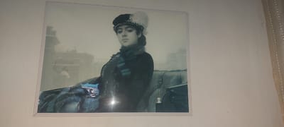 A framed painting of a woman in historical attire is displayed on a wall in a room. The painting depicts a young woman with dark hair, wearing a black cap adorned with white feathers, and a dark coat with a fur collar. She has a green scarf tied around her neck. The background of the painting is a muted, atmospheric scene of buildings and a carriage. The painting itself appears to be behind glass, as reflections are visible on its surface. The wall is a plain, light-colored surface. The room's environment suggests an indoor setting, possibly a home or an art gallery. The specific location in the city of Kandyagash, Kazakhstan is not discernible from the image itself.