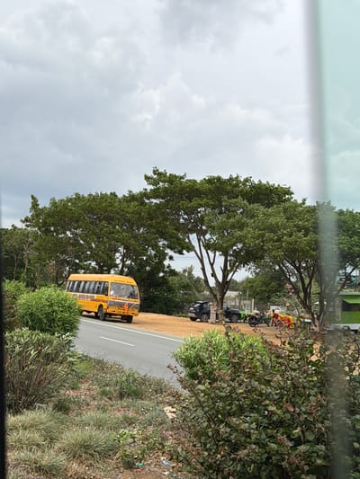 In the city of Addakal, India, a yellow school bus is parked on the side of a road. The bus has the words "SCHOOL BUS" written on its side. Several trees are visible in the background, and the sky is overcast. There are also two cars, a motorcycle, and some people standing near the trees. The scene appears to be a roadside area with vegetation and a dirt patch.