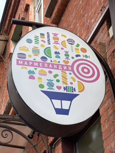 Here's a description of the image:
The image depicts a round, illuminated sign for a confectionery store, mounted on a building's exterior. The sign features a white background adorned with colorful, whimsical candy illustrations and a stylized house shape. The name of the store, "Mармеландия", is printed vertically in pink letters. The sign is attached to the building with a dark metal bracket. The building has brickwork visible in the background. Based on information in other search results, it is possible that this scene is located in Akhieli, Georgia.