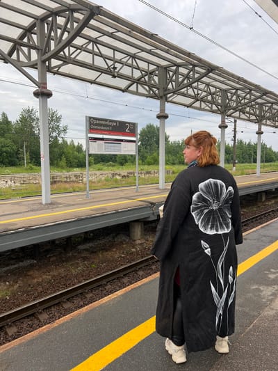 A woman with red hair stands on a train platform in Lomonosov, Russia, facing away from the camera. She is wearing a long, black kimono-style coat with a large white poppy design on the back and white sneakers. The platform has a yellow line marking the edge and a train track runs alongside it. In the background, there are trees and a cloudy sky. A sign with "Ораниенбаум" (Oranienbaum) and train schedule information is visible on the platform.