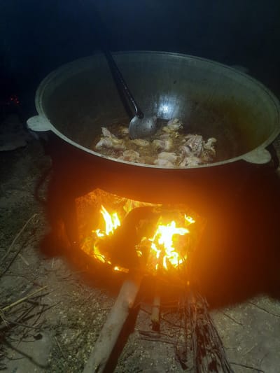 In Восеабад, Tajikistan, an outdoor cooking scene unfolds at night. A large, cast-iron cauldron, likely a kazan, is positioned over a blazing fire built within a makeshift brick or clay oven. The fire casts an intense orange glow upwards, illuminating the contents of the cauldron. Inside the cauldron, pieces of meat, possibly chicken or lamb, are frying in hot oil, with a slotted spoon resting on the edge, ready for use. The surrounding ground appears to be dirt and sparse dry brush, indicating a rural or open-air setting. The darkness of the night contrasts sharply with the bright flames and the cooking activity.