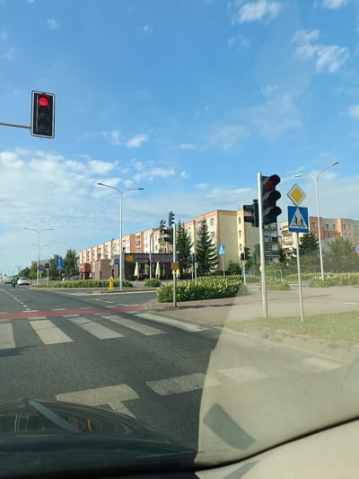 This media file shows a street scene in Ciechanów, Poland, likely during the daytime. The sky is a bright blue with scattered white clouds, suggesting pleasant weather. The scene is dominated by a road with a marked pedestrian crossing. Several traffic lights are visible, with one prominently displaying a red light, indicating that traffic should stop.
In the background, there is a row of apartment buildings with orange and beige facades. In front of these buildings, there is a commercial area with several businesses. One establishment has a sign that says "PIZZERIA" and "DIAVOLO", suggesting a pizza restaurant. Other businesses appear to have awnings and outdoor seating areas.
Streetlights are also present, and there are several trees and bushes lining the road and sidewalks. A few cars are visible on the road, and a person in a green shirt is crossing the street, presumably using the pedestrian crossing. There are also several road signs, including a diamond-shaped yellow sign and a blue sign with a bicycle and pedestrian symbol. A black car, possibly the one from which the photo was taken, is partially visible in the foreground, reflecting the sky and surrounding environment on its windshield. The overall impression is a typical urban streetscape on a clear day.
