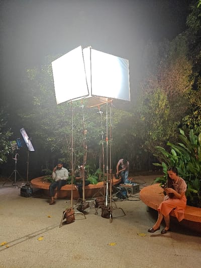 The scene depicts an outdoor film or photography shoot setup at night, located in Kalyan-Dombivli, India. Artificial illumination from multiple sources brightens the central area, creating an atmospheric haze.
In the foreground, a prominent lighting rig features two large, rectangular softboxes emitting bright white light, mounted on multiple chrome light stands, secured by brown sandbags. To the left, a smaller LED panel light on a stand is also active.
Four individuals are present. On the right, a female with long dark hair, dressed in a light-colored top and an orange skirt with black flat shoes, sits on an curved, terracotta-colored stone bench. She is looking down intently at a small, dark object held in her hands. To her left, slightly behind the main lighting rig, a male wearing a dark t-shirt and jeans is bent over, engaged with various pieces of equipment, including cables and power units, on the paved ground.
Further left, on another section of the same terracotta-colored stone bench, a male with a beard, wearing a light collared shirt and dark trousers, is seated and holding a mobile phone to his right ear. Another male is partially visible seated next to him.
The ground is paved with a light-colored material, showing some scattered yellow leaves. The environment is lush, with numerous green trees and large-leafed plants forming the background and surrounding the benches, consistent with a park or garden setting. The sky is dark, indicating nighttime.
