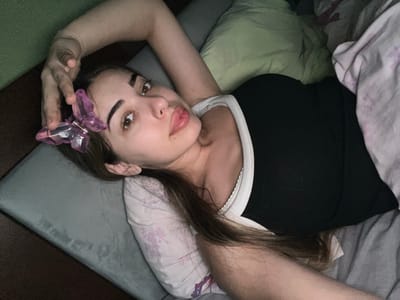 A young woman with long brown hair is lying in bed, looking up at the camera. She is wearing a black tank top with a white trim. A pink butterfly hair clip is attached to her hair. She has a diamond stud earring in her left ear. The room appears to be a bedroom, with pillows and blankets visible. The lighting is dim, suggesting it might be nighttime or early morning. The location is not specified in the image.