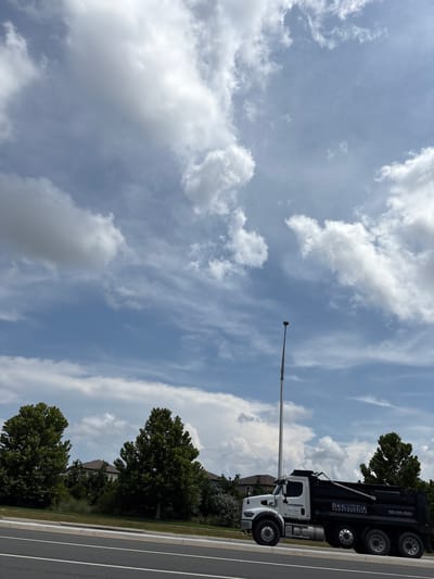 This is a vertical shot of a dump truck with the text "MOGANIER0 CONSTRUCTION" visible on its side. The truck is white and black, and it appears to be parked on the side of a road. The road has white lane markings. Behind the truck, there are trees and houses. The sky is blue with white clouds, suggesting it is daytime and the weather is fair. A tall pole with a light fixture extends upwards from the ground, partially obscuring the sky. The scene is set in Parrish, United States. No people are visible in the image.