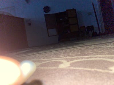 The image shows an interior scene, possibly a room, in low light conditions. There is a shelf unit with multiple compartments against a white wall. A carpet or rug with a patterned design is partially visible in the upper left corner of the image. A bright light source is present in the upper right corner, obscuring details. The room appears to have furniture and other items. It is not possible to confirm the location in Sabo, Nigeria, or any specific activities within the room.
