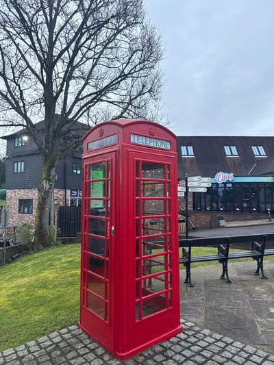A K6-style bright red telephone box stands prominently on cobblestone paving, positioned slightly right of the image center. It features clear glass panels with white mullions, a light blue "TELEPHONE" sign with white lettering above the door, and a crown emblem on its upper panels. The door displays a "PULL" handle. To the telephone box's left, a manicured green grass slope ascends towards a large, bare deciduous tree. Behind the tree, a multi-story building with dark grey/black timber cladding and visible brickwork on its lower section features multiple rectangular windows. A "PRO SHOP" sign is partially visible on a dark fence line in front of this building. To the right of the telephone box, the cobblestone paving transitions into a larger area of grey flagstones, where two black metal benches with wooden slats are situated. Further in the background, across a paved path or road, is a building with a dark pitched roof and several evenly spaced skylights. The ground floor of this building features large glass windows, displaying a prominent sign for "Olie's Waffles & Coffee" in light blue and pink lettering, with "WAFFLES & COFFEE" visible below. A wooden directional signpost with multiple white arrows is located to the left of the "Olie's" building. The sky is overcast and grey. The scene is located at Old Mill Lane in Sheet, Petersfield, within the district of East Hampshire, United Kingdom.
