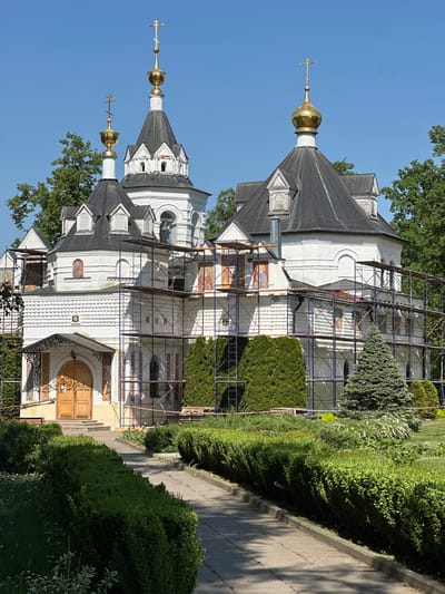 The image captures the architectural details of a Russian Orthodox church, possibly located in Махра, Russia. The church features a white exterior with intricate brickwork and black roofs. Golden onion domes with crosses are visible atop the structure. Scaffolding surrounds parts of the church, suggesting ongoing maintenance or restoration work. Lush greenery, including bushes and trees, frames the building, while a pathway runs alongside it. The scene is bathed in sunlight under a clear blue sky.