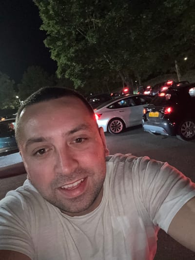 A close-up, slightly angled self-portrait of a smiling man wearing a white t-shirt. In the background, cars are lined up on a street at night. A tree is visible on the left side of the image. One car has a license plate "RJ1 ECH". Based on the license plate, the location could be Borough of Spelthorne, United Kingdom.