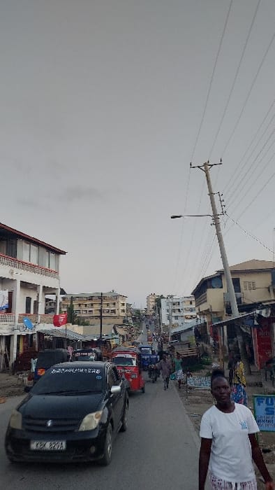 This image depicts an active urban street scene within Magogoni ward, Kenya, under an overcast sky. The view is looking uphill along a paved road.
In the immediate foreground on the right, an adult female of African descent, wearing a white short-sleeved t-shirt and light-colored patterned shorts or skirt, walks towards the viewer. Her hair is dark and styled upwards. Behind her, a blue sign with white text is partially visible next to a makeshift roadside structure with a corrugated iron roof.
On the left side of the road, a black Toyota Ist sedan (Kenyan registration KBX 829A) is positioned, facing away from the viewer. Its front bumper and windshield are visible, with some text or decals across the top of the windshield. The vehicle appears to be part of a queue of traffic.
Further up the street, traffic is dense, including at least two red and blue auto-rickshaws (tuk-tuks), motorcycles, and other vehicles. Numerous pedestrians are navigating the street, walking on the shoulders and within the traffic lanes.
Along the left side of the road, a prominent multi-story building with a white facade and multiple balconies with dark railings is visible. Laundry items are hanging on one of the balconies. A blue satellite dish is affixed to the lower portion of this building. Red banners or flags are also present near this structure. Further uphill, various other multi-story buildings line the street.
On the right side of the road, a tall, light-colored wooden utility pole stands prominently, supporting multiple overhead power lines and a street light fixture. Adjacent to and behind this pole are more buildings, some appearing to be older or less developed, with various small businesses or stalls at street level.
The road recedes into the distance, ascending and curving slightly to the left, with more buildings visible in the background, indicative of a densely populated urban area. The sky is uniformly grey, suggesting an overcast day.