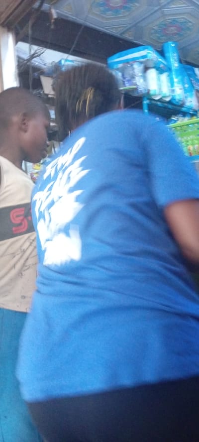 In Kwachiri, Nigeria, a medium shot captures two individuals standing in what appears to be a store. On the left, a person with dark skin and short hair is seen from the side, wearing a light-colored tank top with a dark band and a red "S" emblem. To the right and slightly in front, another person, whose back is to the camera, is wearing a bright blue t-shirt with a white graphic design on the back. The background reveals shelves stocked with packaged goods, including blue and white plastic packaging, suggesting a retail environment. The ceiling above has a decorative tiled pattern. The overall lighting suggests an indoor setting, possibly during the day.