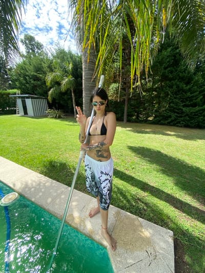 A person stands poolside, cleaning with a long-handled tool. They wear sunglasses, a black bikini top, and patterned white shorts. Tattoos adorn their arms and torso. The pool's edge is stone, with bright green water. Lush green grass extends beyond the pool, shaded by trees. A small metal structure is visible in the background near the grass. The photo is taken in daylight.