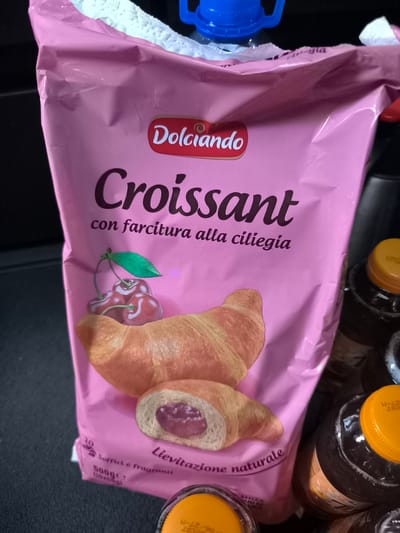 A close-up shot shows a pink package of Dolciando croissants with cherry filling. The package features a large illustration of croissants and cherries. Text on the package reads "Dolciando Croissant con farcitura alla ciliegia" and "Lievitazione naturale". Other text visible includes "500ge (10x50g)" and "Soffici e fragranti". In the background, several dark bottles with yellow caps are visible, some with partially obscured text, possibly beverages. The lighting suggests an indoor setting. The overall impression is a product shot focused on food items, likely for a retail or marketing purpose.