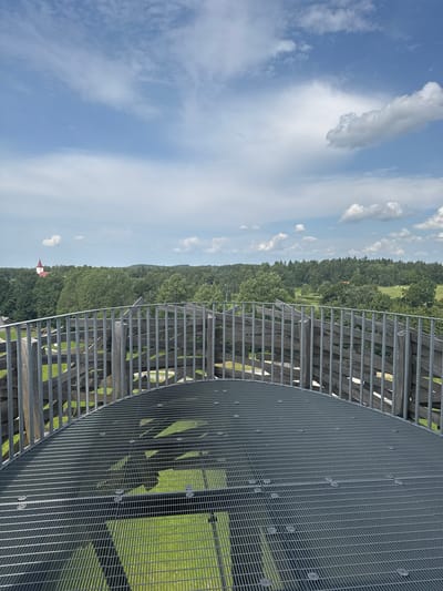 An eye-level perspective view from the top of the Rõuge observation tower in Rõuge, Estonia, shows a curved metal grate platform in the foreground, with vertical wooden and metal railings forming a circular enclosure around it. Through the railings, a panoramic view of a lush green landscape unfolds, characterized by dense, dark green trees stretching towards the horizon. In the distance, a white building with a red, pointed roof, likely a church, is visible amidst the foliage. The sky above is a bright blue, punctuated by scattered white, fluffy clouds. The overall scene depicts a clear, daytime view of a natural environment from an elevated vantage point.